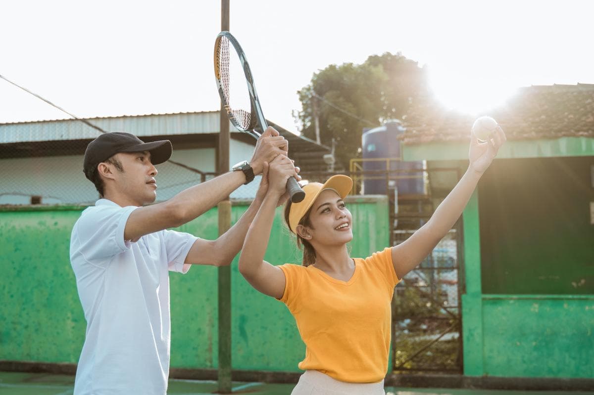 Seorang pemain tenis wanita sedang berlatih servis bersama pelatih pria di lapangan tenis.